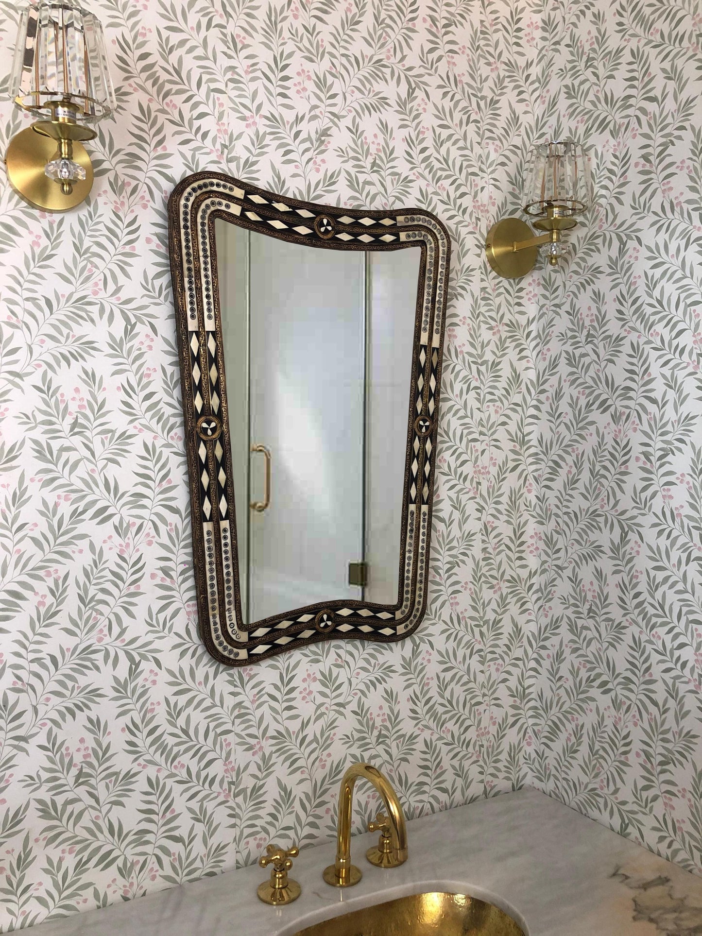 Mirror with brass faucet and floral wallpaper.