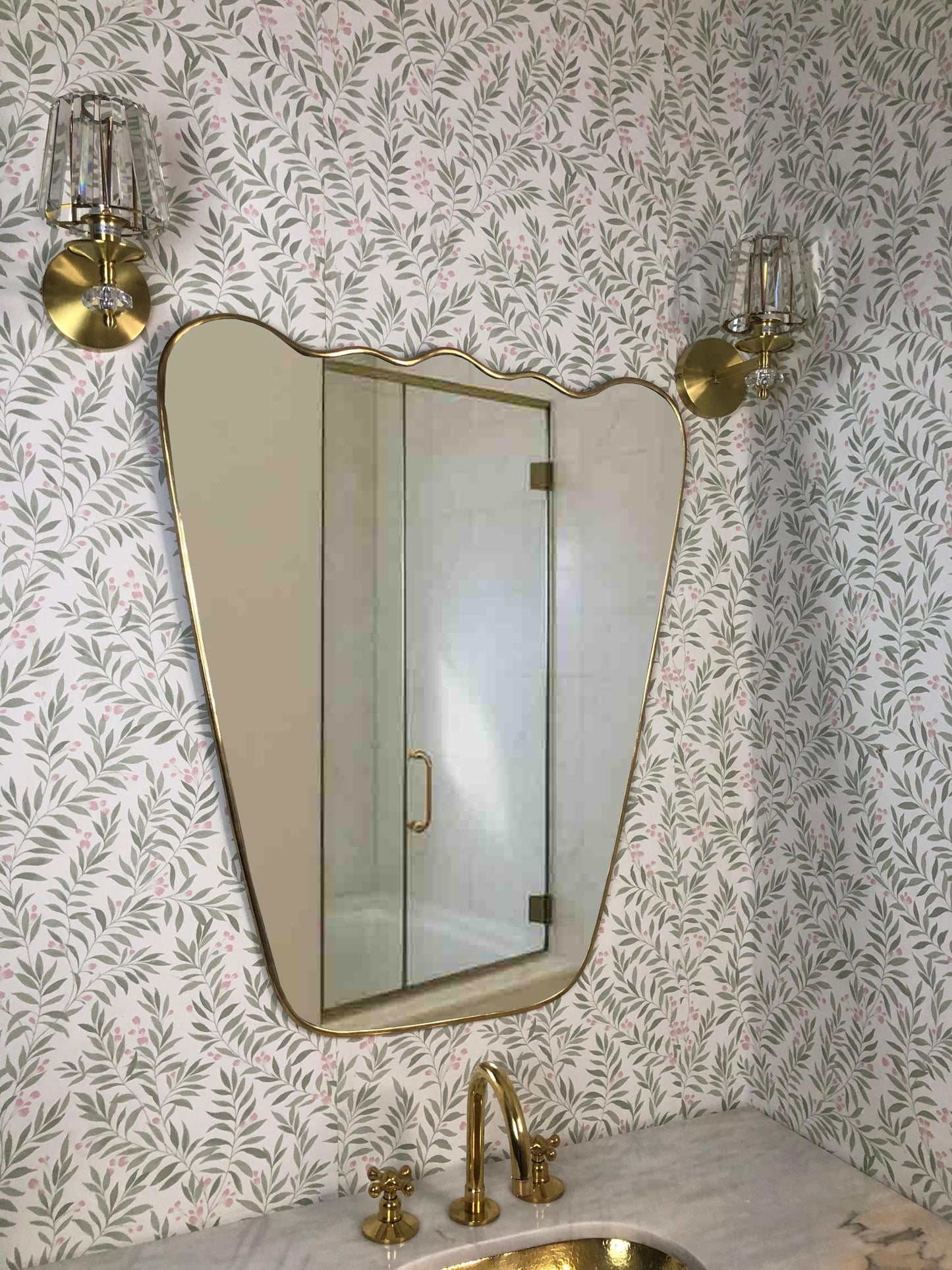 Wavy brass-framed mirror above bathroom sink with floral wallpaper.