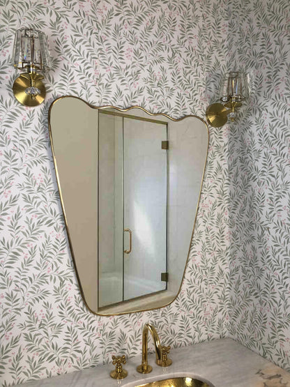 Wavy brass-framed mirror above bathroom sink with floral wallpaper.
