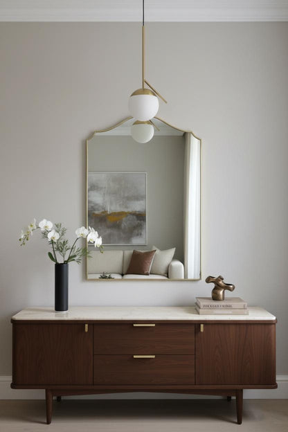Brass arched mirror above wooden dresser, decorated with orchid flowers, books, and modern pendant light.