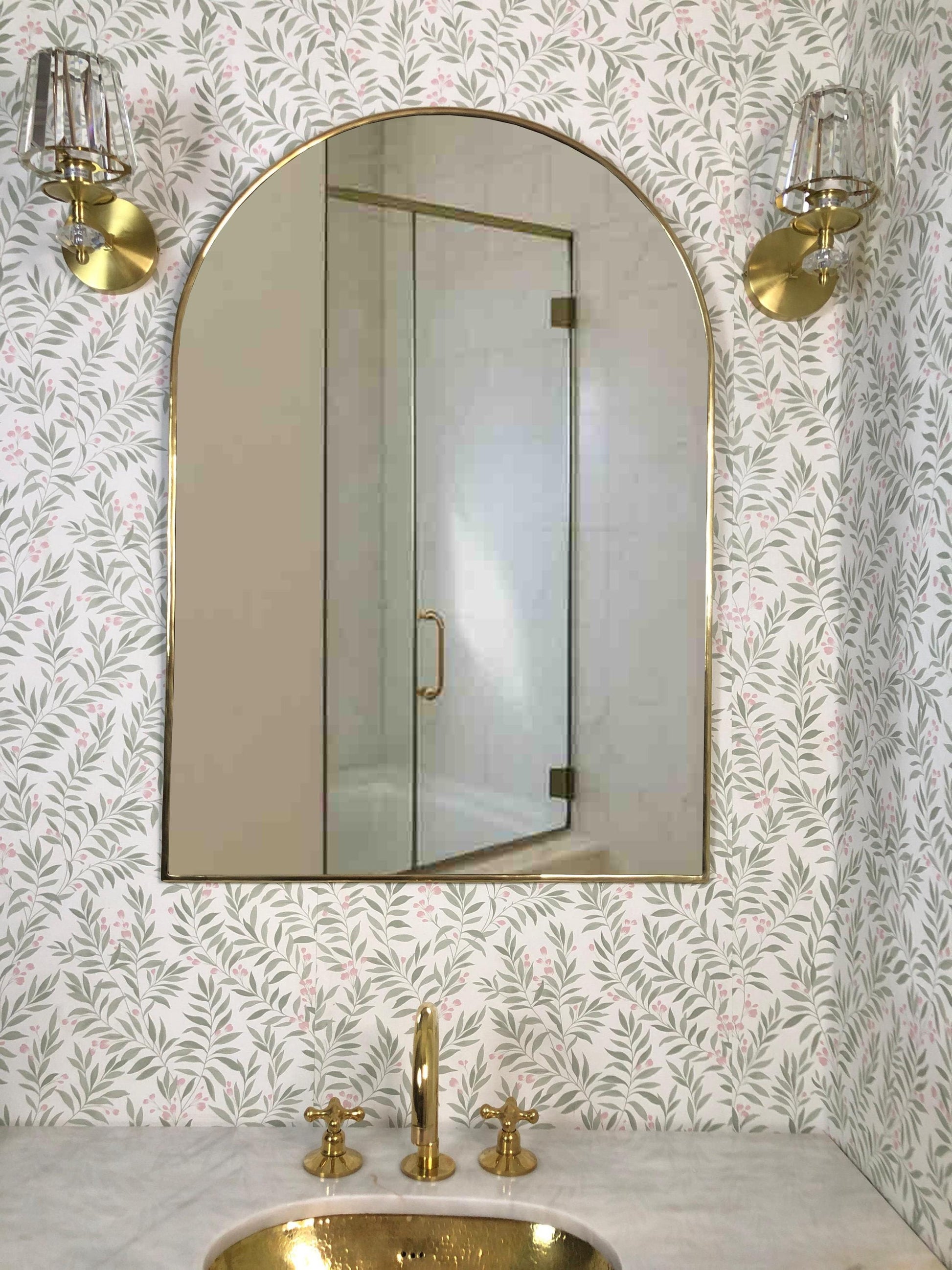 Arched brass wall mirror above a bathroom sink with brass fixtures.