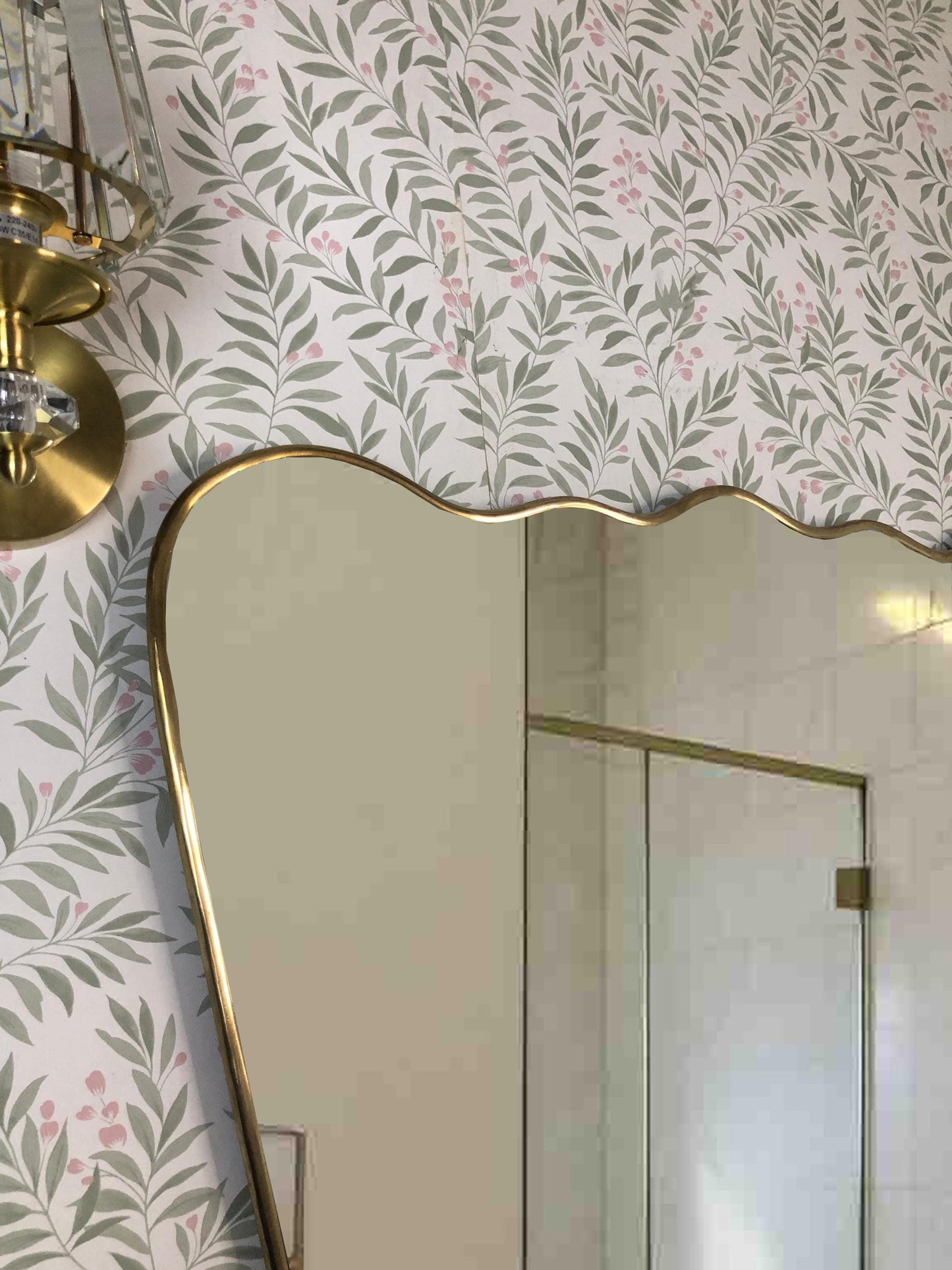 Upper corner of brass mirror near brass wall sconce on floral wallpaper.