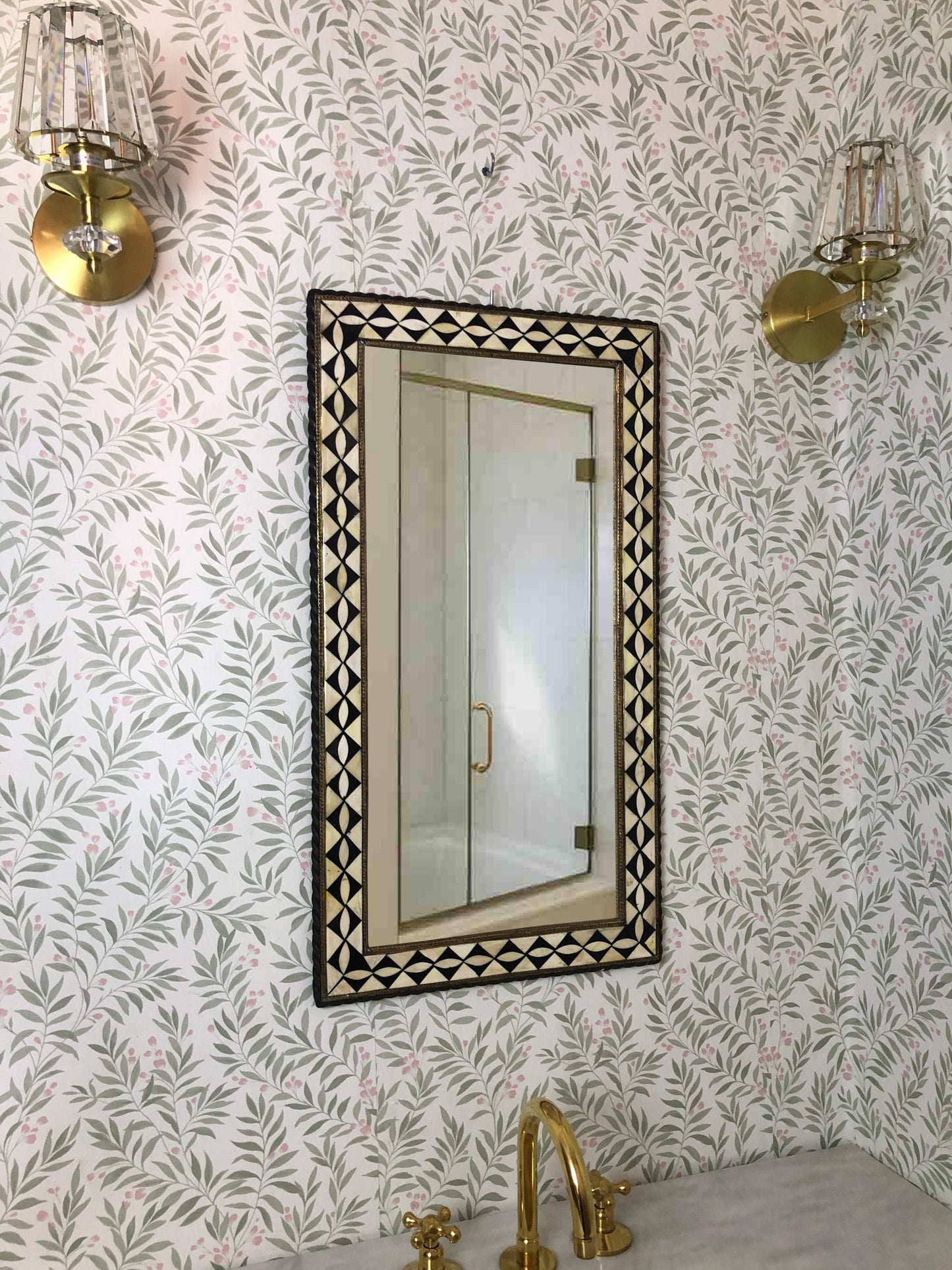 Bathroom view with brass fixtures, floral wallpaper, and geometric framed mirror.