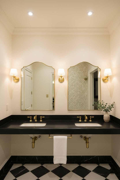 Pair of brass framed arched mirrors above double vanity with black marble counter, brass faucets, and wall sconces