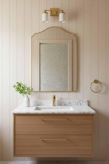Arched brass framed mirror above light wood vanity with marble top, gold faucet, and small vase with greenery