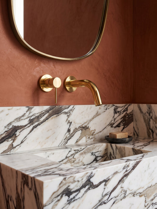 Modern unlacquered brass wall-mounted bathroom faucet installed above a marble integrated sink with warm terracotta walls.