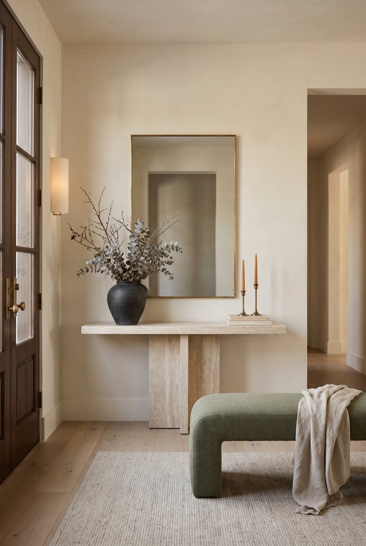 Slim rectangular brass mirror above a stone console with a dark vase, leafy branches, and taper candles.