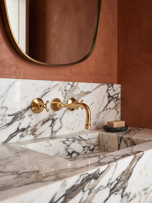 Close-up of a polished unlacquered brass wall-mounted faucet installed on dramatic marble backsplash.