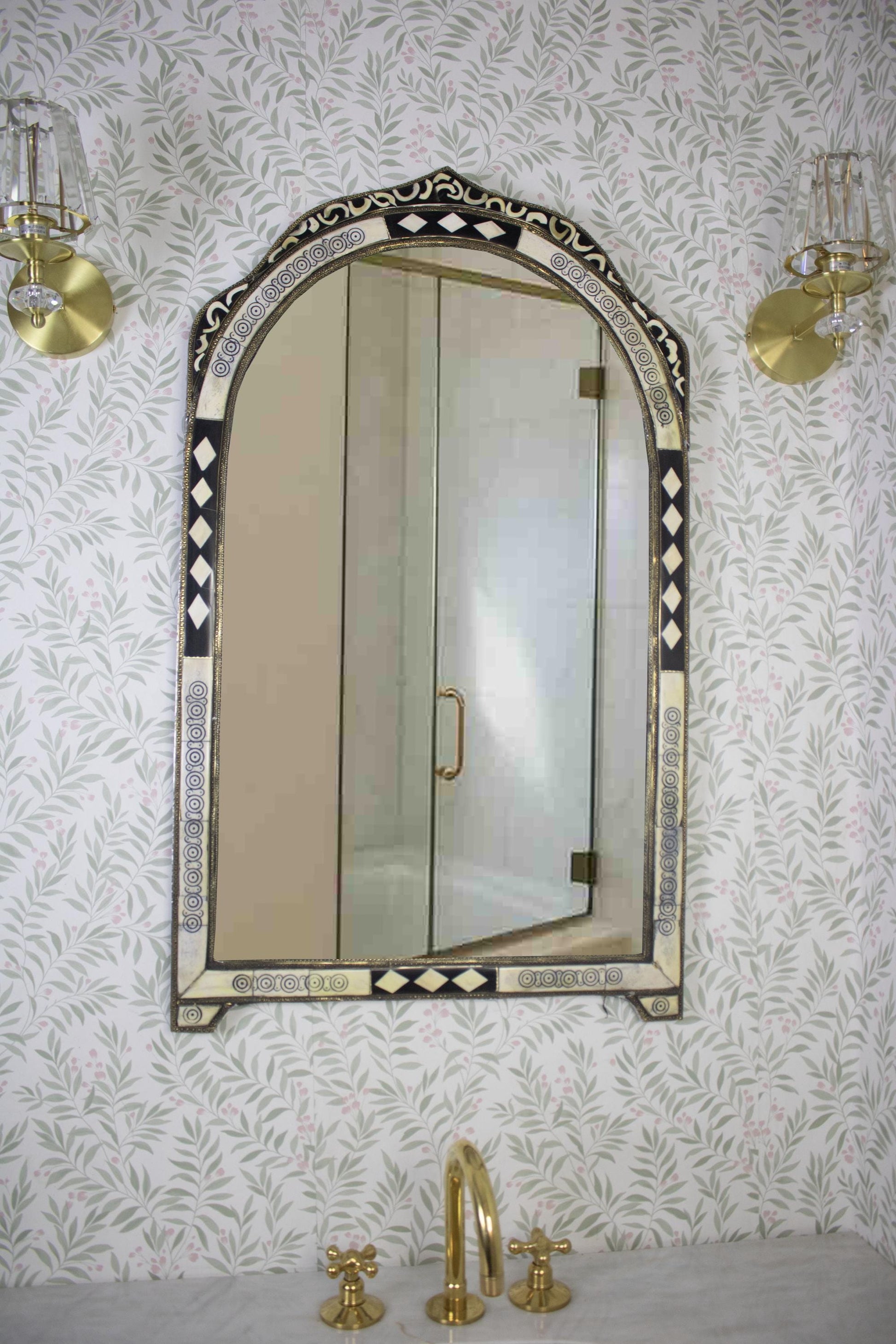 Arched Moroccan mirror above a sink with brass faucet and floral wallpaper.