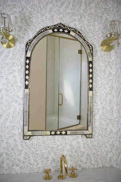 Arched Moroccan mirror above a sink with brass faucet and floral wallpaper.
