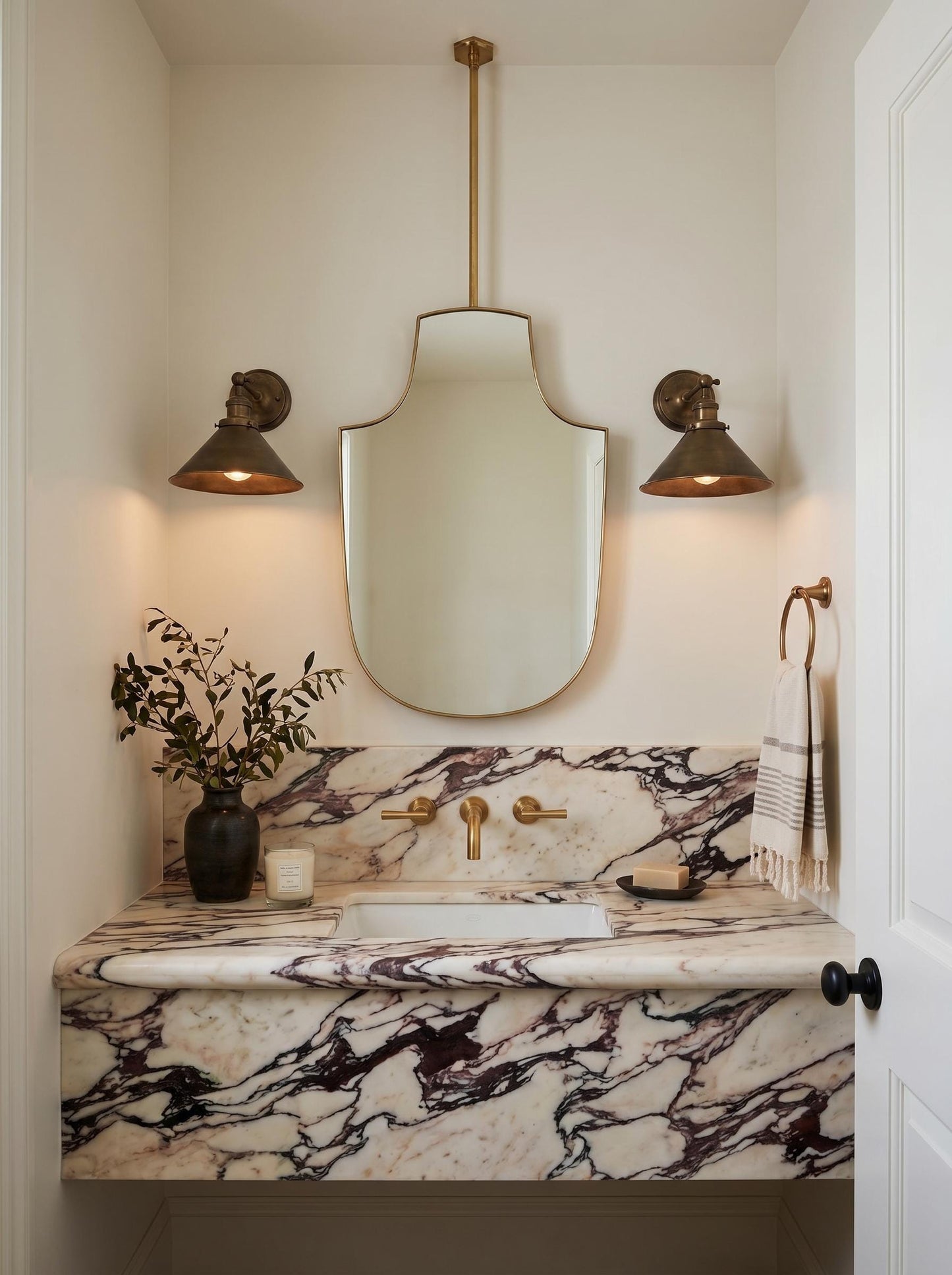 Suspended shield brass mirror styled over marble vanity with brass sconces