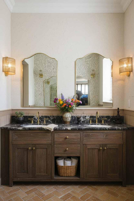 Pair of brass framed mirrors above rustic dark wood vanity with marble counter, wicker sconces, and floral arrangement